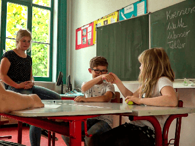 Screenshot: Offener Kanal Magdeburg Zwei Kinder, die sich die Hände schütteln, beobachtet von einer Schulsozialarbeiterin